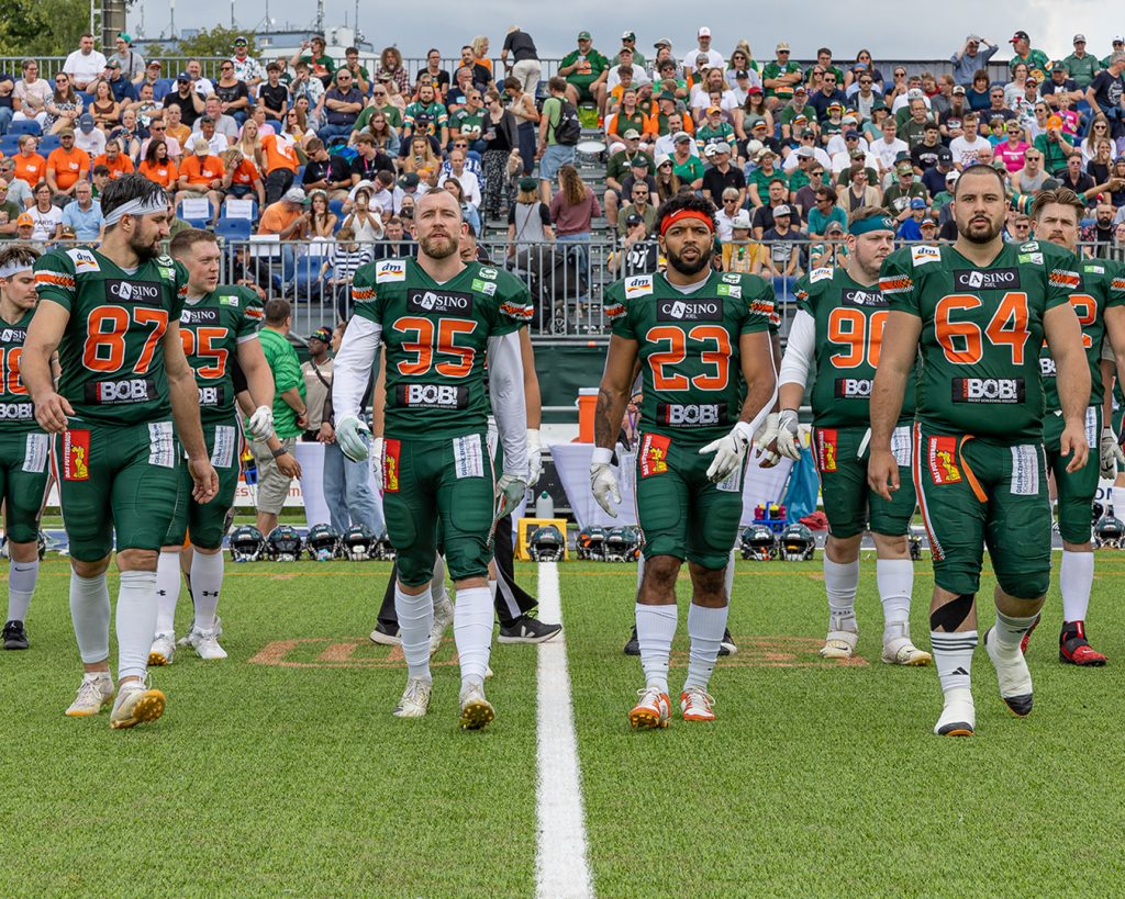 Auf dem Foto sieht man die vier Team Captains 2024 von vorn zum Münzwurf auf das Feld gehen. Hinter geht der Rest des Teams in einer Linie und dahinter stehen die Fans auf der Tribüne. Die vier Captains sind von links nach rechts Benedikt Englmann, Bodo Bilitewski, CJ Davis, Kevin von Kroge.