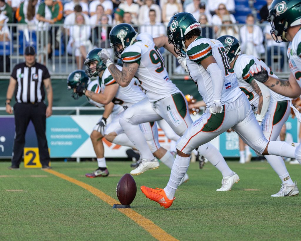 Cobian Adjei-Freeman führt den Kickoff zum White Out Game 2023 am 02.09.23 gegen Potsdam aus. Er und die die anderen Spieler hinter ihm im Bild tragen die komplett weiße Uniform der Hurricanes. Im Hintergrund auf der Tribüne sieht man auch die Zuschauer in weißer Kleidung.